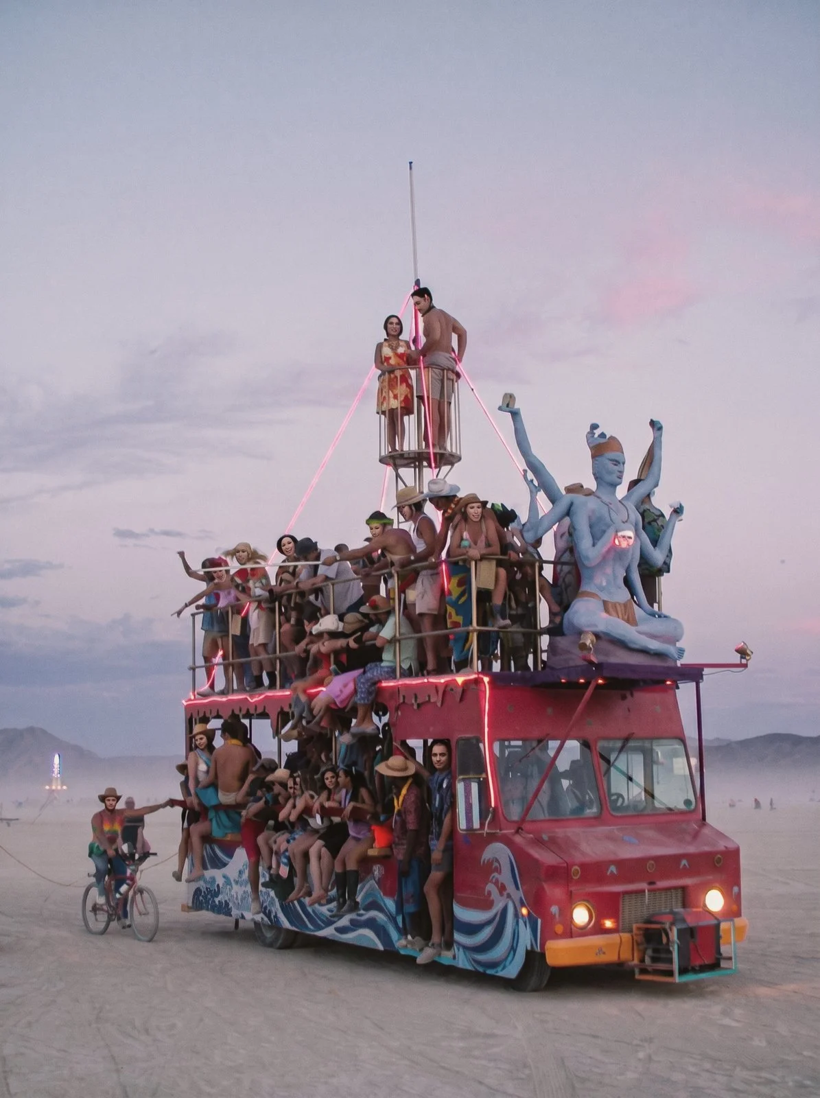 Red art car bus full of dancers with a blue goddess statue on the desert playa at dusk