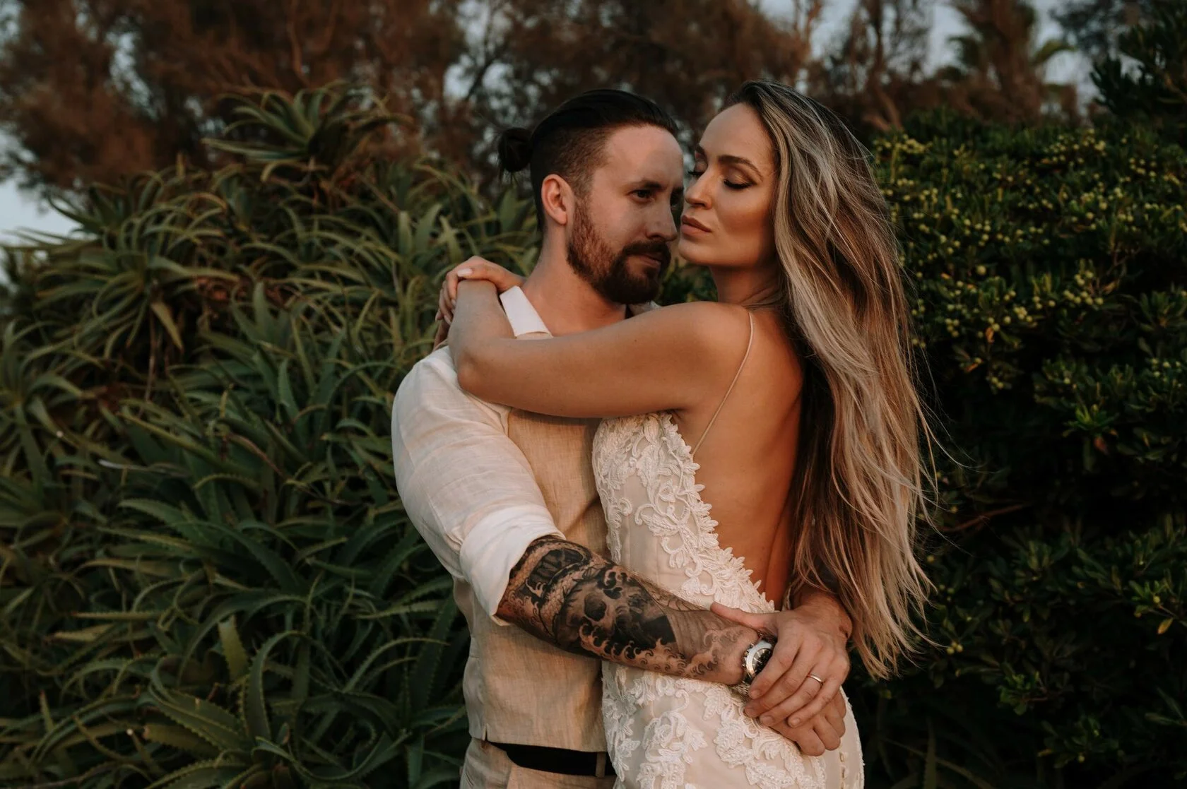 Groom hugs the bride among green bushes at sunset