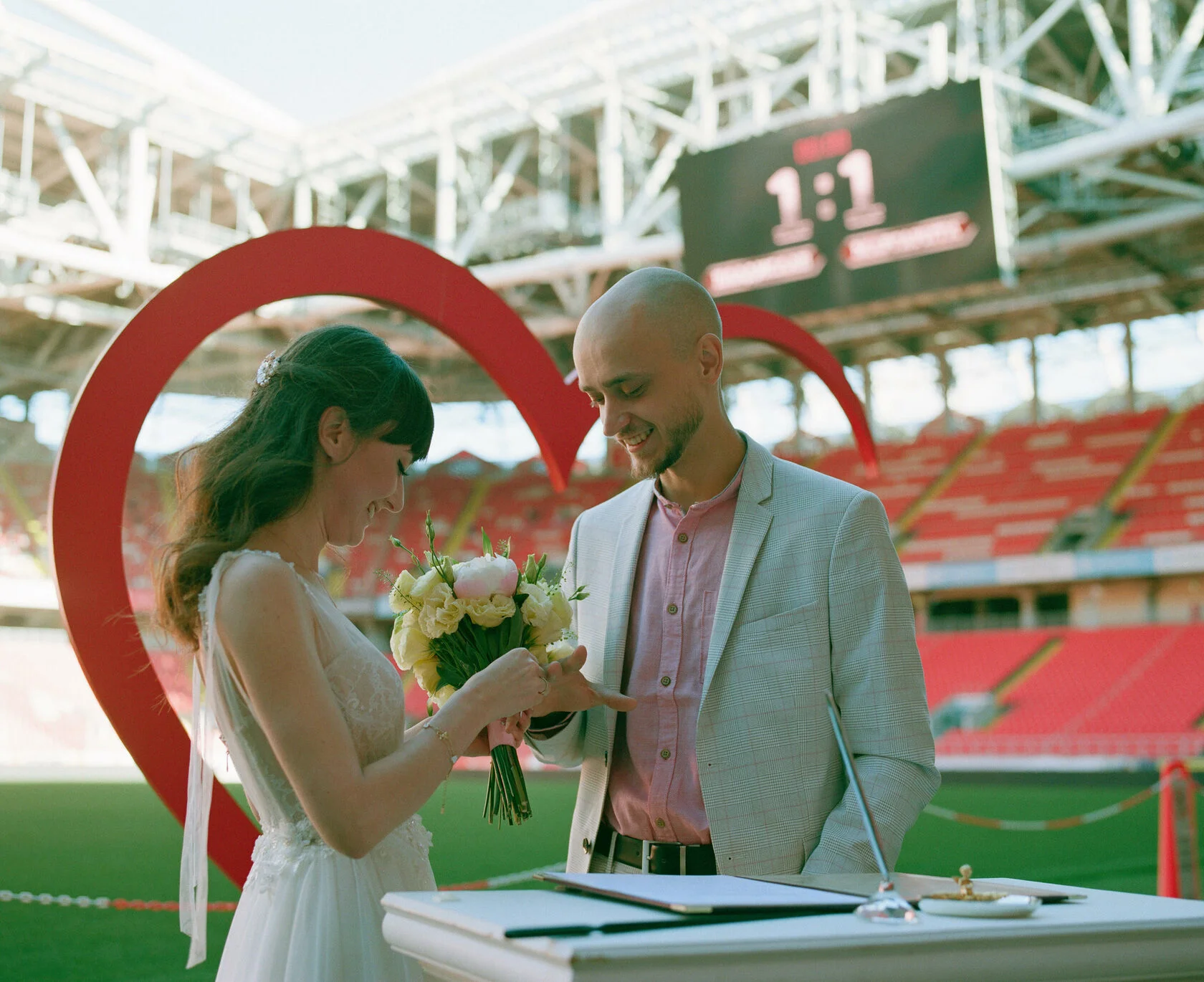 Bride and groom sign at a table in front of a big red heart on the stadium pitch