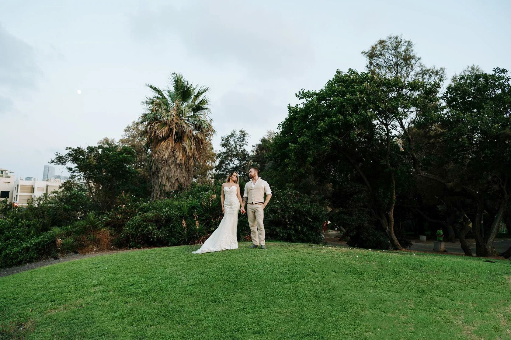 Bride and groom hold hands on a green hill under palm trees in Tel Aviv