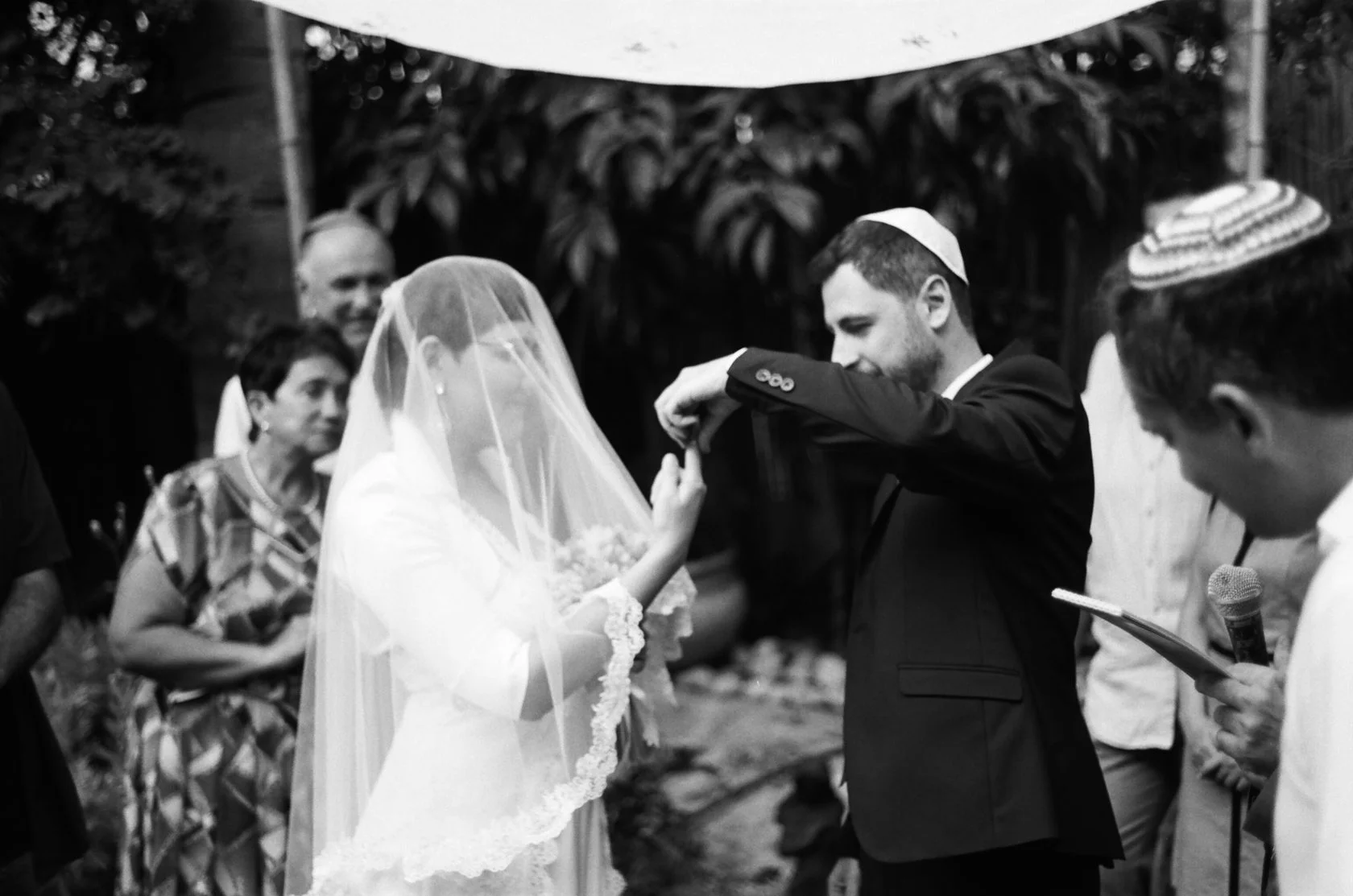 Black-and-white film photo: the groom lifts the bride's veil under the chuppah
